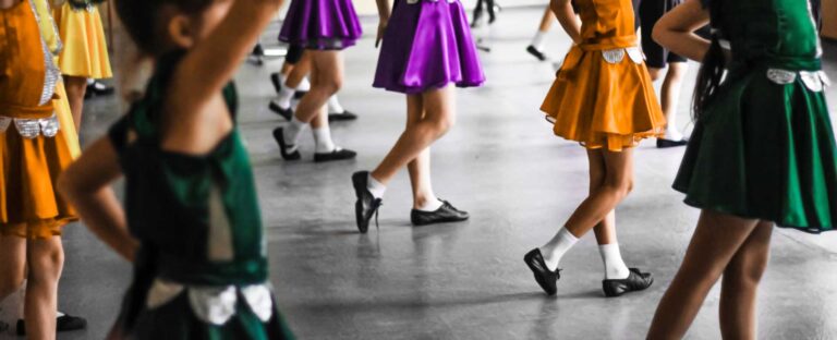Děti v barevných sukýnkách a tanečních botách tančí v řadě na podlaze tanečního sálu. Některé mají paže nah